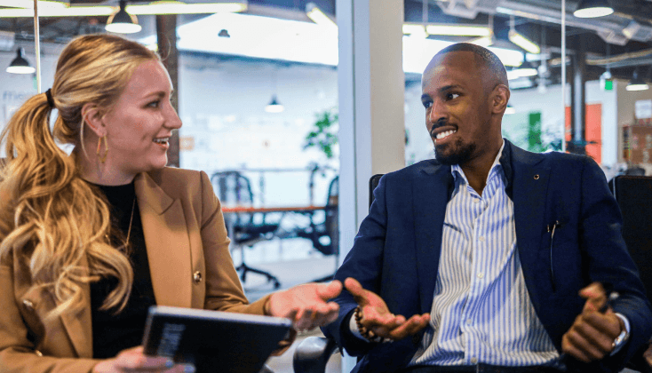 A woman and a man engage in conversation in a modern office setting, both smiling and gesturing.
